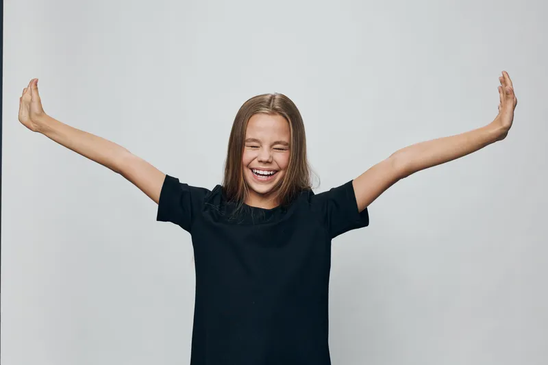Girl with long hair smiling and stretching arms wide wearing black t shirt on light background. Happy child, emotion expression, casual clothing and carefree gesture for joy and freedom.