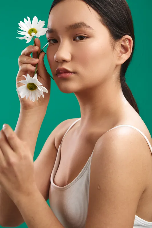 asian woman presents a calm studio portrait, softly holding daisies against a vivid green backdrop, capturing a fresh beauty, gentle mood, and natural light that highlights graceful features and
