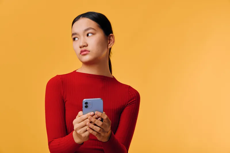 Young woman with smartphone in hands looking away on orange background. Female model in red sweater holding mobile phone for communication and social media connection.