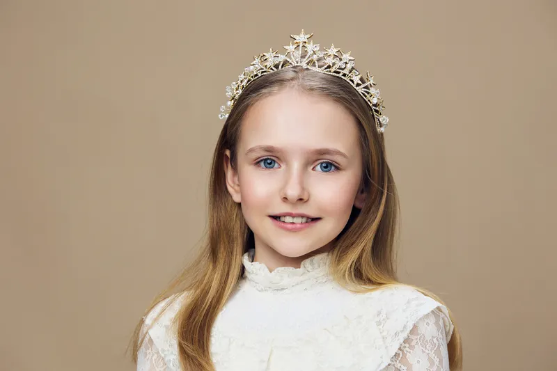 Portrait of a young girl with a crown, wearing a lace dress and smiling, set against a beige background, evoking innocence and elegance, perfect for children s fashion