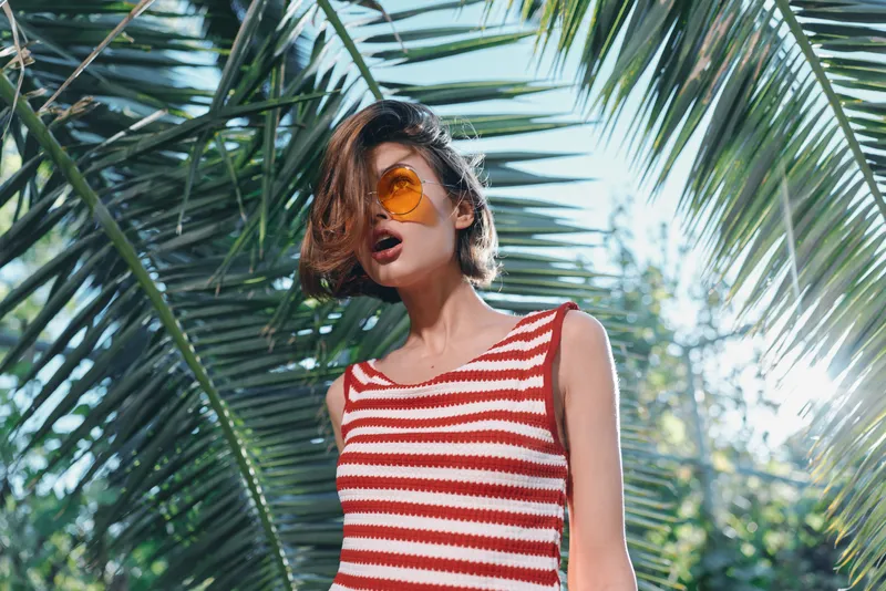 A stylish woman wearing a red and white striped sleeveless top stands in a sunny tropical setting. She wears round sunglasses, gazes off camera, and radiates a relaxed, playful vibe.