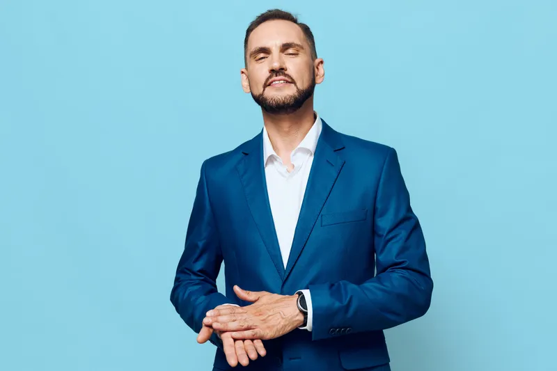 A professional man in a blue suit stands against a solid colored backdrop, exuding calm confidence and strong business poise in a studio portrait.