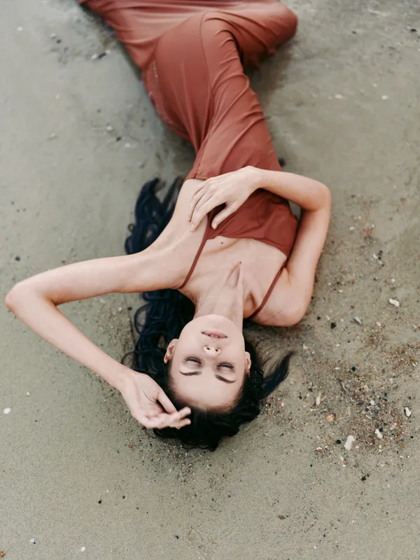 A serene portrait of a woman lying on a sandy beach in an orange dress, eyes closed, hair spread, and a calm, graceful pose illuminated by soft sunlight.