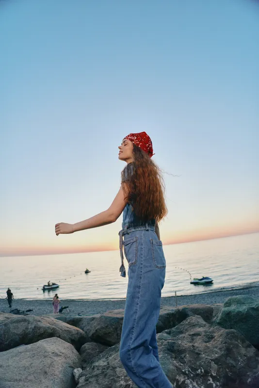 young woman with red bandana standing on rocks by the sea during sunset, wearing denim overalls and enjoying calm coastal atmosphere