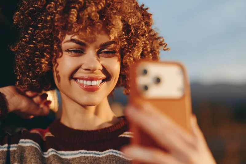 Woman taking selfie outdoors, curly hair framing her face, bright smile and relaxed eyes, holding a smartphone in hand, warm autumn light, lifestyle moment, blurred background, scenic