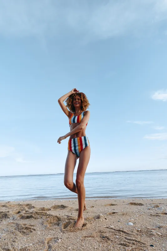 Young woman with curly hair enjoys a sunny day on the beach wearing a colorful striped bikini, standing barefoot on the sand with a joyful expression under a clear blue sky.