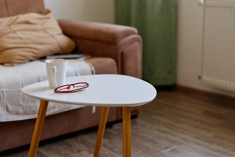 Calm living room scene with a small round table, a cup and mug resting on its surface, a soft sofa and cushions in the background, warm interior tones highlighting a cozy home mood