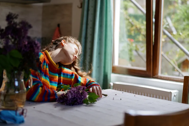 Young woman wearing a rainbow striped sweater rests her head on a table beside a window, with flowers in the foreground. Cozy domestic scene showing comfort, relaxation, and home life, soft natural