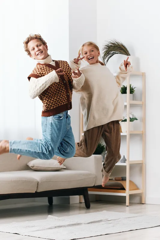 Two joyful children jump in mid air indoors, wearing cozy sweaters and jeans, capturing a moment of playful energy and carefree happiness in a bright living room.