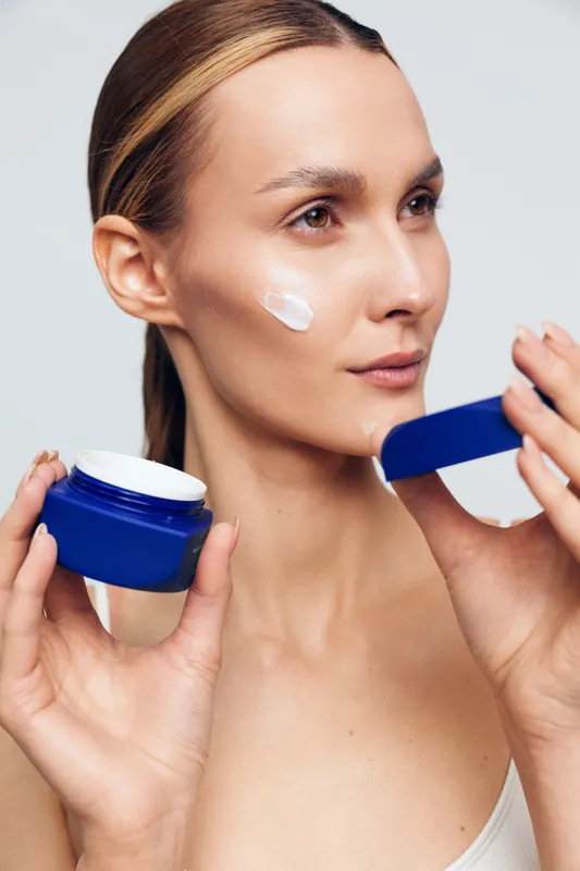 Woman applying skincare cream on her face, holding blue jar and spreading white lotion with fingers, calm beauty routine, concept of self care and wellness today