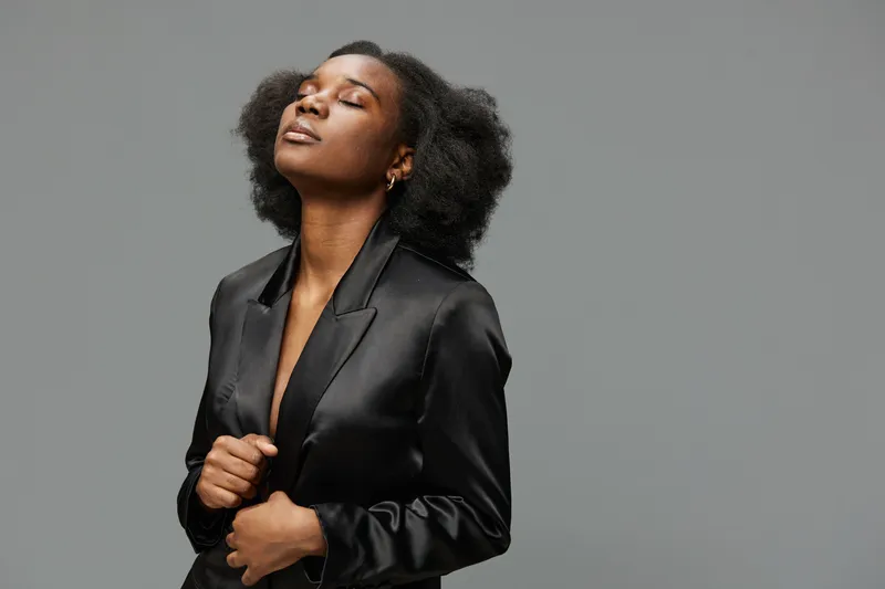 Confident young woman with natural hair in a black blazer, exuding calmness and self assuredness against a soft gray background, ideal for professional and fashion themes