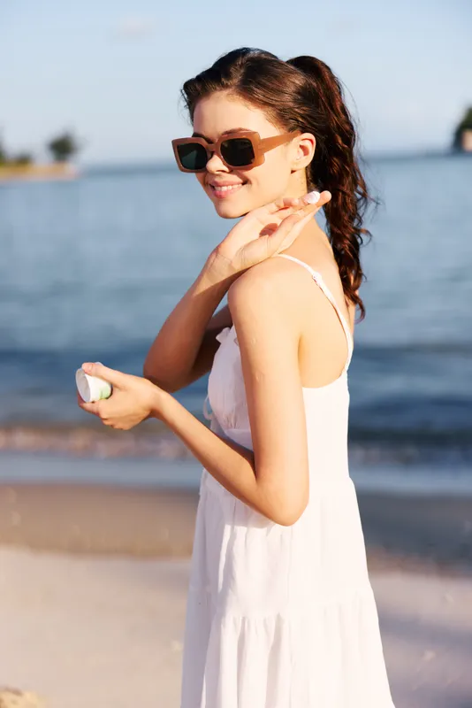 Young woman in a white sundress and sunglasses stands on a sunny beach, smiling while holding a seashell, with calm sea and sky in the blurred background.