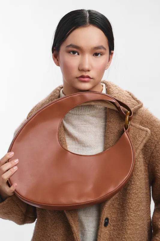Asian woman holding a circular brown leather bag while wearing a cozy sweater and coat, presenting a chic, contemporary fashion moment with an understated sense of elegance.