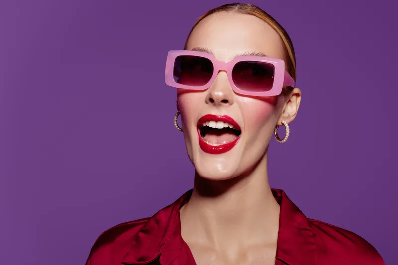 Stylish woman with pink sunglasses and red lipstick smiles confidently against purple background. Fashion concept with bold makeup, accessories, and vibrant colors for modern style.