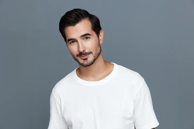 Smiling young man dressed in a casual white t-shirt stands against a solid grey background. His confident expression captures a sense of modern lifestyle and approachability.
