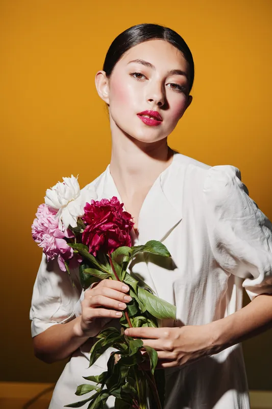 Stylish woman in white dress holding pink and red flowers, posing against warm yellow backdrop. Beauty and fashion creative concept with polished makeup and elegant expression.
