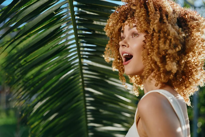 smiling woman with curly hair enjoys sunny tropical nature, surrounded by lush green palm leaves in bright daylight, radiating happiness and natural beauty.