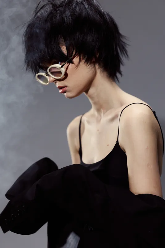 Stylish young Asian woman with short black hair wearing white sunglasses and a black outfit in moody lighting.