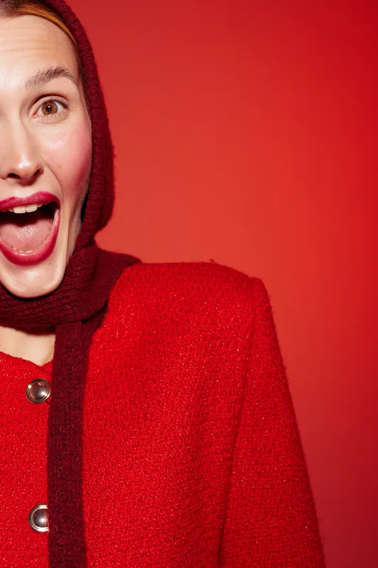 Excited woman in red coat and scarf smiles widely with open mouth against red background. Concept of happiness, fashion, winter clothing, and vibrant emotions in studio lighting.