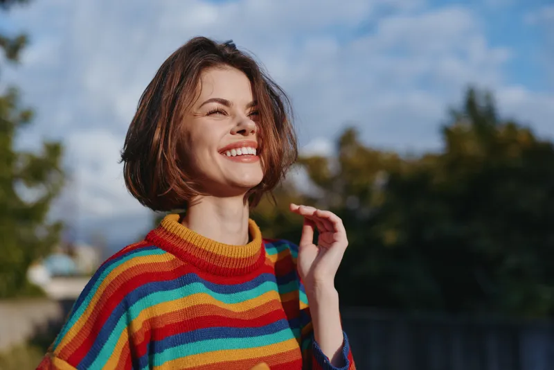 Woman, smile, portrait, colorful sweater, outdoors, happy, female, young — closeup of a joyful woman wearing a rainbow striped sweater in natural light, casual short hair and relaxed expression.
