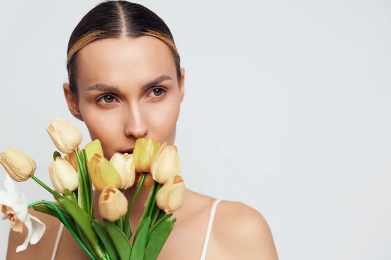 A young woman with natural makeup holds a bouquet of yellow tulips and white orchids close to her face against a plain light background. Concept of spring and beauty.