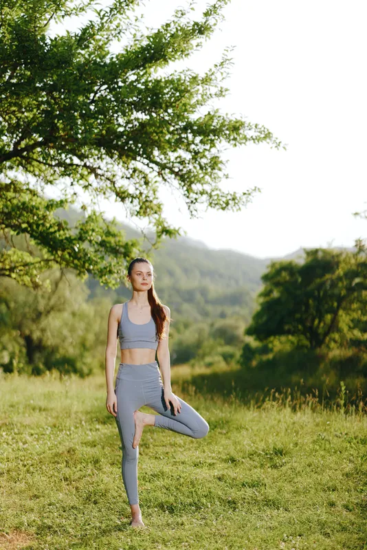 Serene young woman practicing yoga outdoors in a lush green landscape, wearing gray activewear, expressing calmness and balance