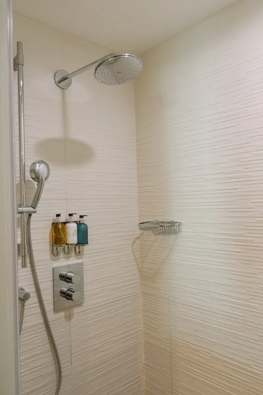 Shower with round overhead head and handheld sprayer in a tiled bathroom, clean chrome fixtures, glass enclosure and wall rack with bath products.