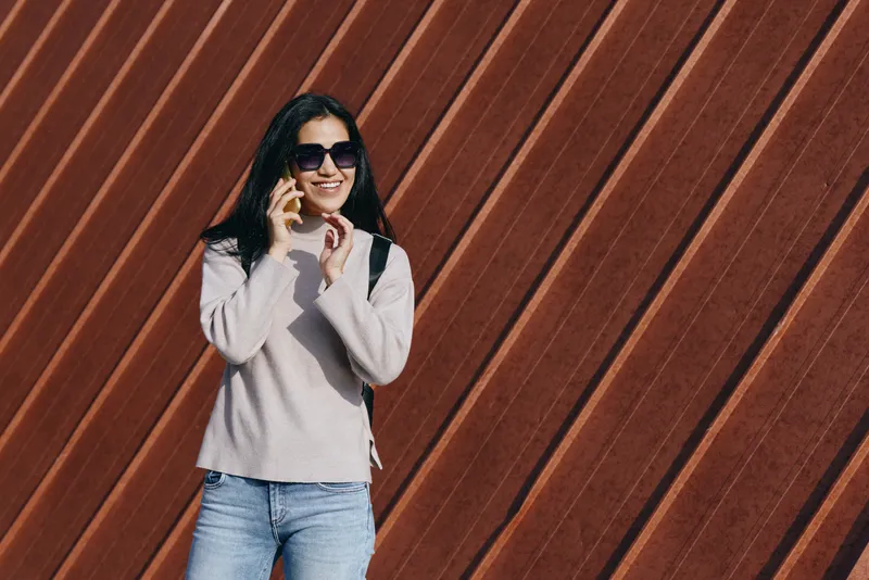 Woman wearing sunglasses talks on a smartphone, dressed in a light sweater and jeans against a diagonal wooden wall, conveying casual urban style and modern communication