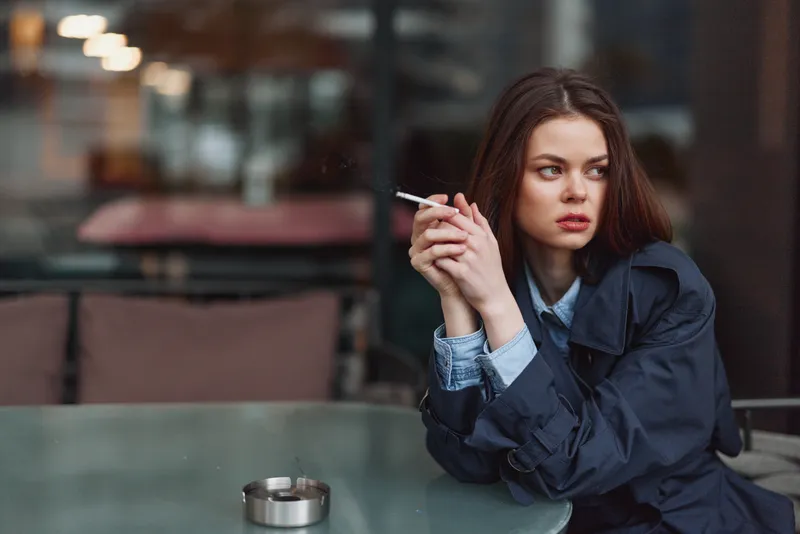 Young woman in dark coat smokes cigarette while sitting alone at outdoor cafe table with ashtray, looking away with thoughtful expression in urban environment.