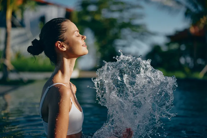 Young woman with brunette hair enjoys a refreshing swim in an outdoor pool while splashing water with her hands on a sunny day, expressing relaxation and joy.