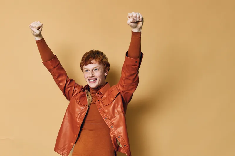 Young man with red hair and freckles wearing a brown leather jacket raising his arms in victory and smiling with excitement on beige background in studio.