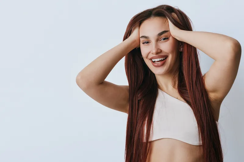 Young woman with long hair smiling and holding head with hands, wearing casual top against plain background. Female model portrait with happy expression and relaxed pose.