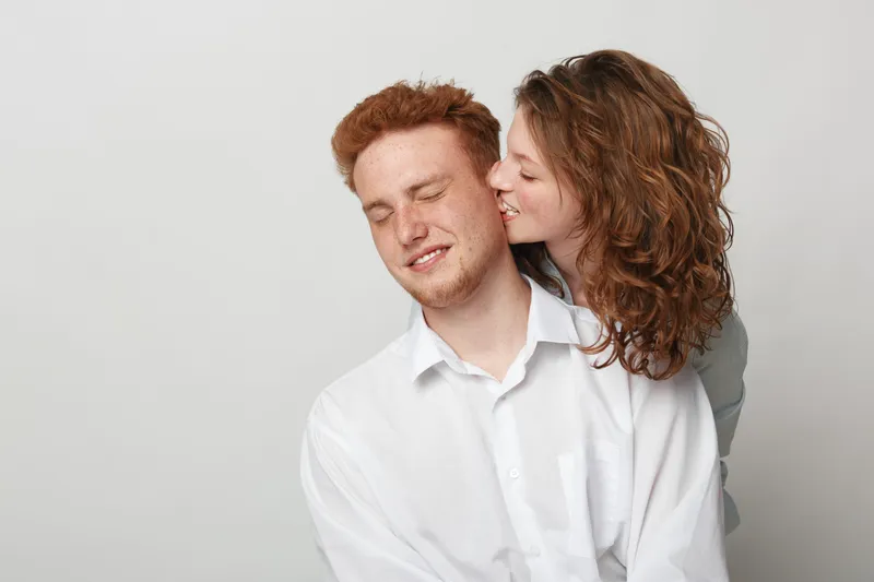 Redhead man with closed eyes smiles while woman with curly hair kisses his cheek. Intimate moment of affection and love between couple on neutral background in casual clothing.
