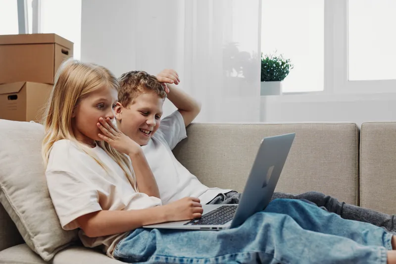 Two happy children sitting on a couch using a laptop together in a bright living room. Concept of technology, digital learning, and playful interaction between siblings.