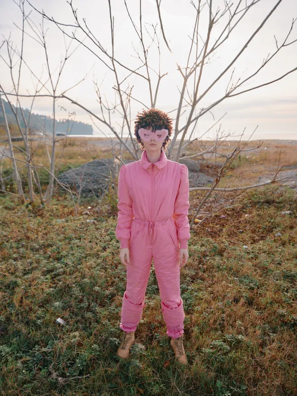Outdoor portrait of a person in a pink jumpsuit standing in a bare field with leafless trees and soft autumn light, displaying retro fashion and quirky style.