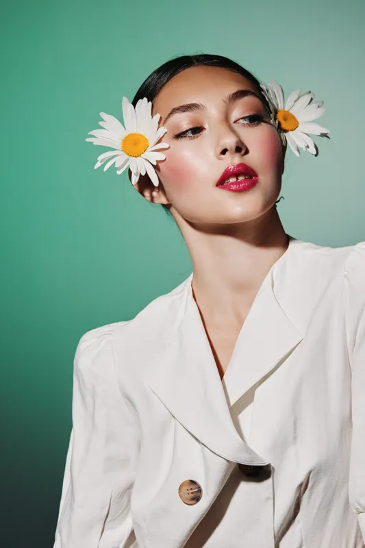 Fashion portrait of a young woman with makeup, red lipstick and white flowers decorating hair. White blazer against green background, beauty and creative fashion concept.