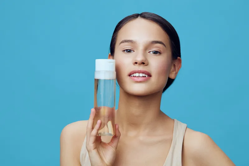 Young woman holding a skincare product with a bright blue background, showcasing a joyful expression and natural beauty, emphasizing healthy skin care and beauty routine