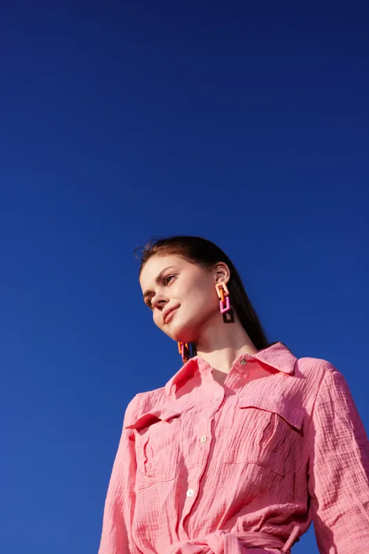 Young woman with long hair in a pink shirt, standing against a clear blue sky, showcasing vibrant fashion and a confident expression ideal for lifestyle photography