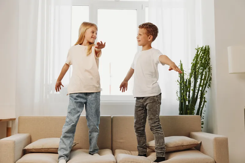 Two happy children jumping on a beige sofa in a bright living room, enjoying playful moments and sibling fun together at home.