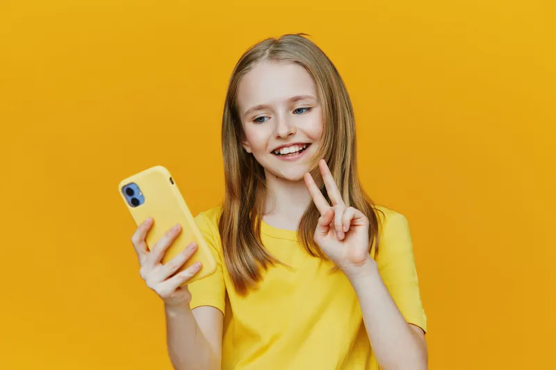 Happy girl with long hair in yellow shirt taking selfie on smartphone against yellow background, showcasing joy and playful emotions in a vibrant setting