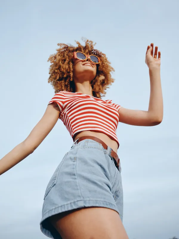 A carefree young woman poses outdoors in a striped crop top and denim shorts, sunglasses, and a sunny breeze, capturing a vibrant moment of confidence and joy.