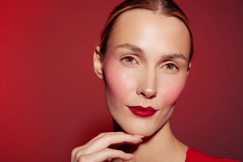 Close-up portrait of a woman with glossy red lips and pink blush on cheeks against red background. Concept of beauty, makeup, skincare, and fashion style.
