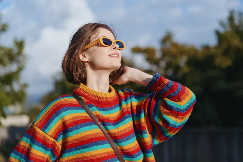 Woman in a colorful striped sweater and yellow sunglasses, smiling and looking up outdoors. Young adult with short brown hair, casual outfit, relaxed travel lifestyle vibe in sunlight.