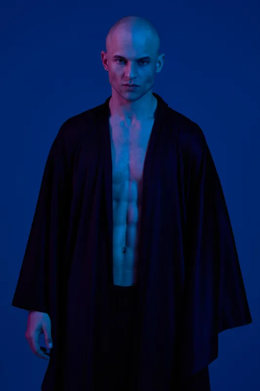 Strong man in a dramatic black robe, standing confidently against a vibrant blue background. The image features a blend of colors highlighting his physique.