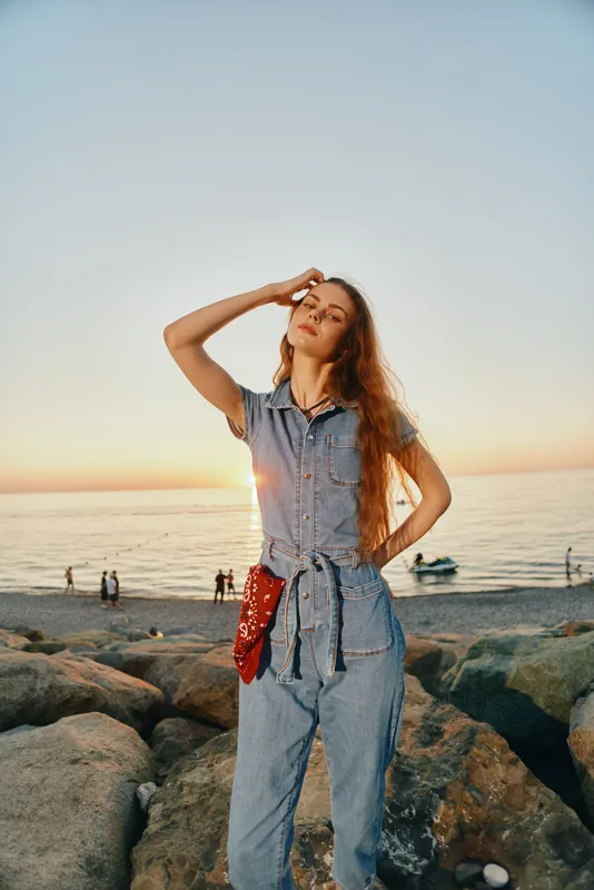 Lifestyle woman in modern boho-western denim street style poses confidently on rocky beach at sunset with film color tones enhancing natural mood and warm atmosphere.