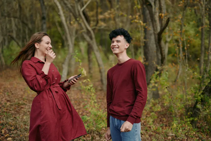 A candid moment conveying authenticity and credibility as two people share a warm exchange in autumn light, dressed in deep red, strolling a forest path.