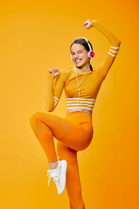 Female fitness dancer in bright orange athletic wear, mid-movement pose with headphones, against a vivid orange background, conveying energy, balance, and dynamic sport vibes for wellness and active