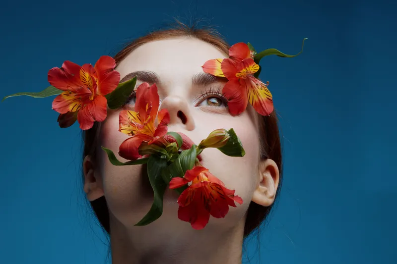 Young woman with flowers on her face, exuding calmness and beauty against a blue background, showcasing vibrant colors and a unique artistic concept