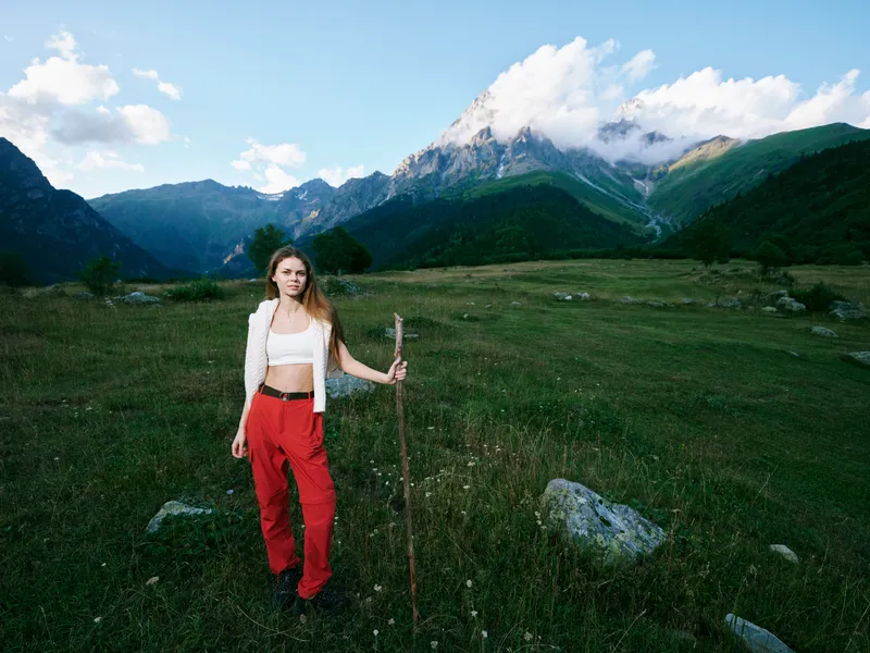 Woman with a wooden staff stands in a green meadow beneath towering mountains, wearing a white jacket and red pants, capturing a tranquil alpine hiking scene amid wildflowers