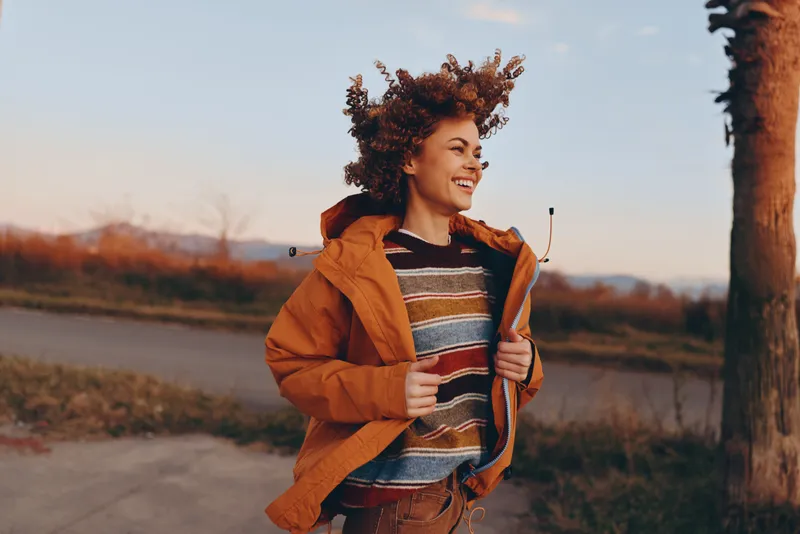Woman outdoors in autumn, orange jacket, smiling while walking on a rural path, soft sunset light, countryside field and trees, lifestyle concept, calm mood